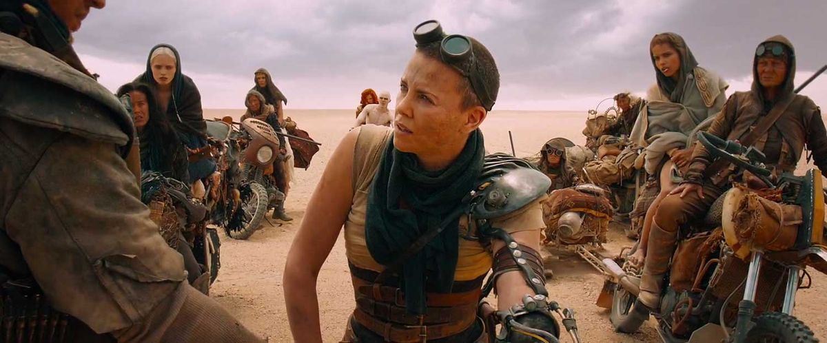 Mad Max Fury Road: A desert dune with a cloudy sky. Over the shoulder shot of Max's shoulder centered on Furiosa, with the Wives and Many Mothers on motorcycles behind her.