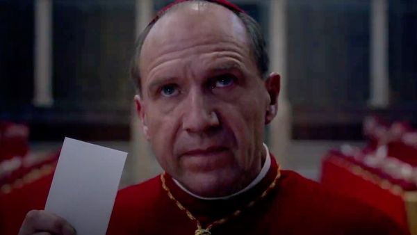 Ralph Fiennes dressed as a Catholic cardinal holds up his ballot to cast a vote for the next pope.