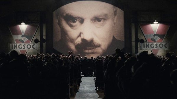 Screencap 1984: A giant image of Big Brother looks down on an assembled crowd. Large INGSOC banners hang on either side of the monitor.