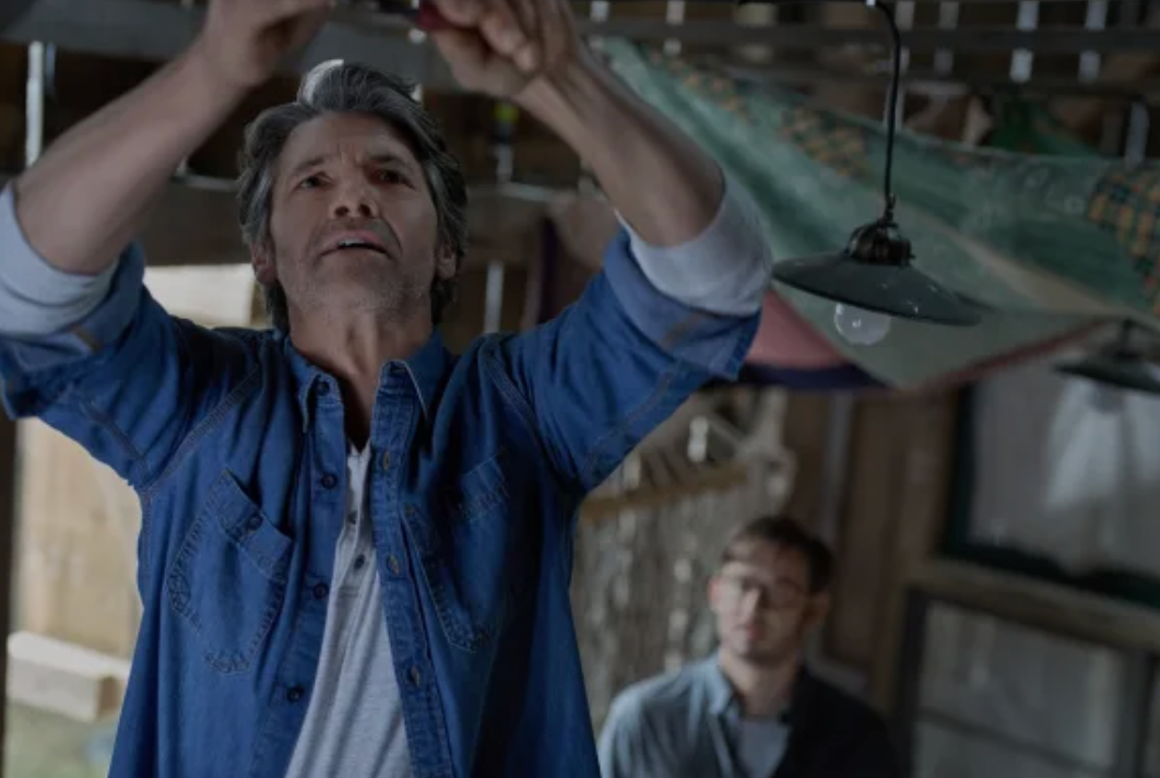 Colton Landry stands in the foreground in a barn repairing a piece of equipment. Eliot Augustine looks on in the background. 