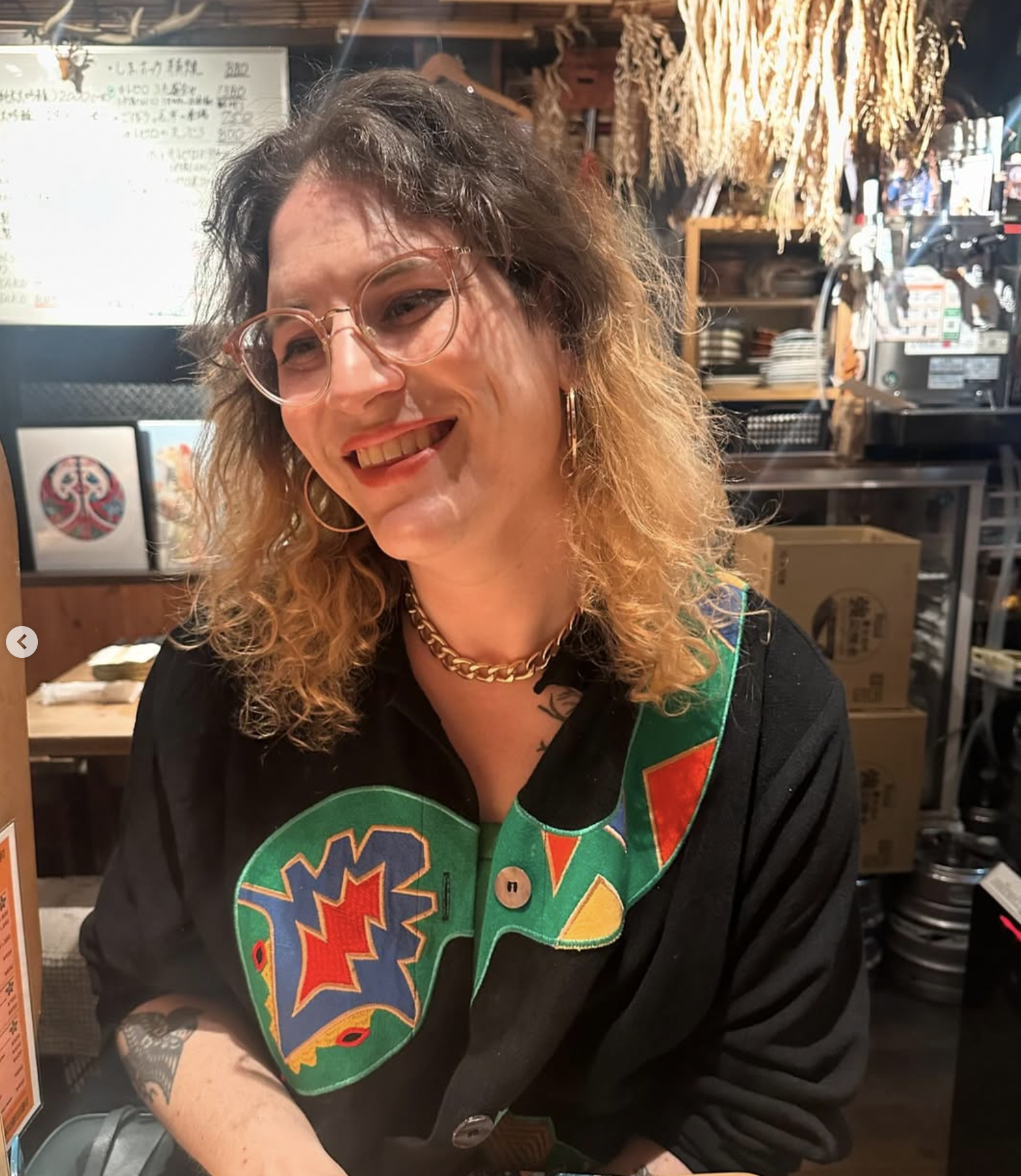 A color photograph of Mattie Lubchansky with curly shoulder-length hair wearing glasses and a colorful patterned top, sitting in a restaurant. She smiles at the camera.
