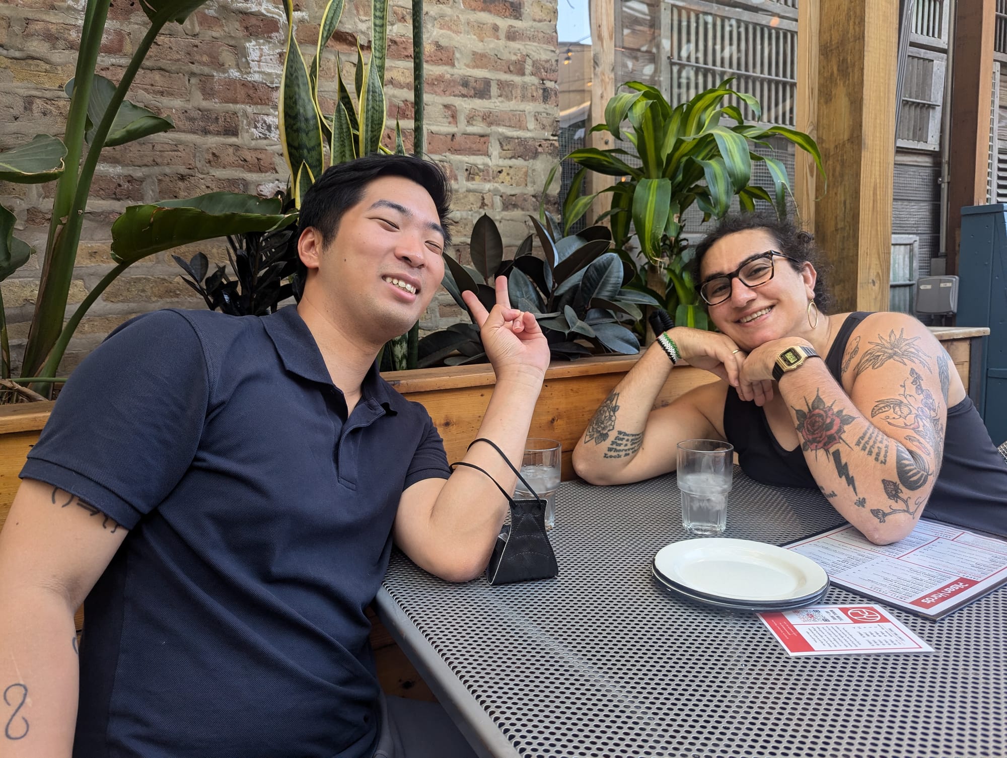 Alex Hanna, smiling, sits outside at a restaurant's patio table with a friend, giving a peace sign.