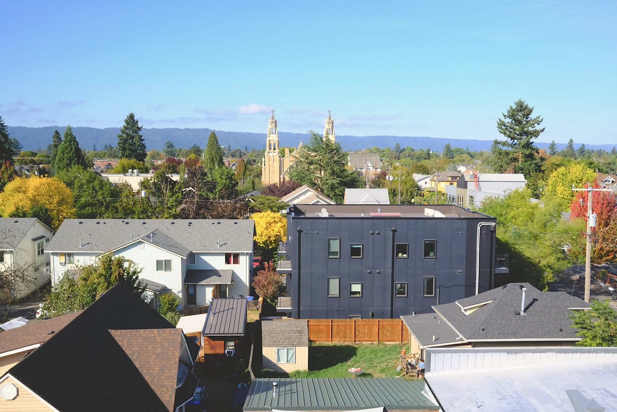 A blue-sky view of the Northeast Portland cityscape including the spires of a cathedral, tiny houses in backyards, and the West Hills beyond.