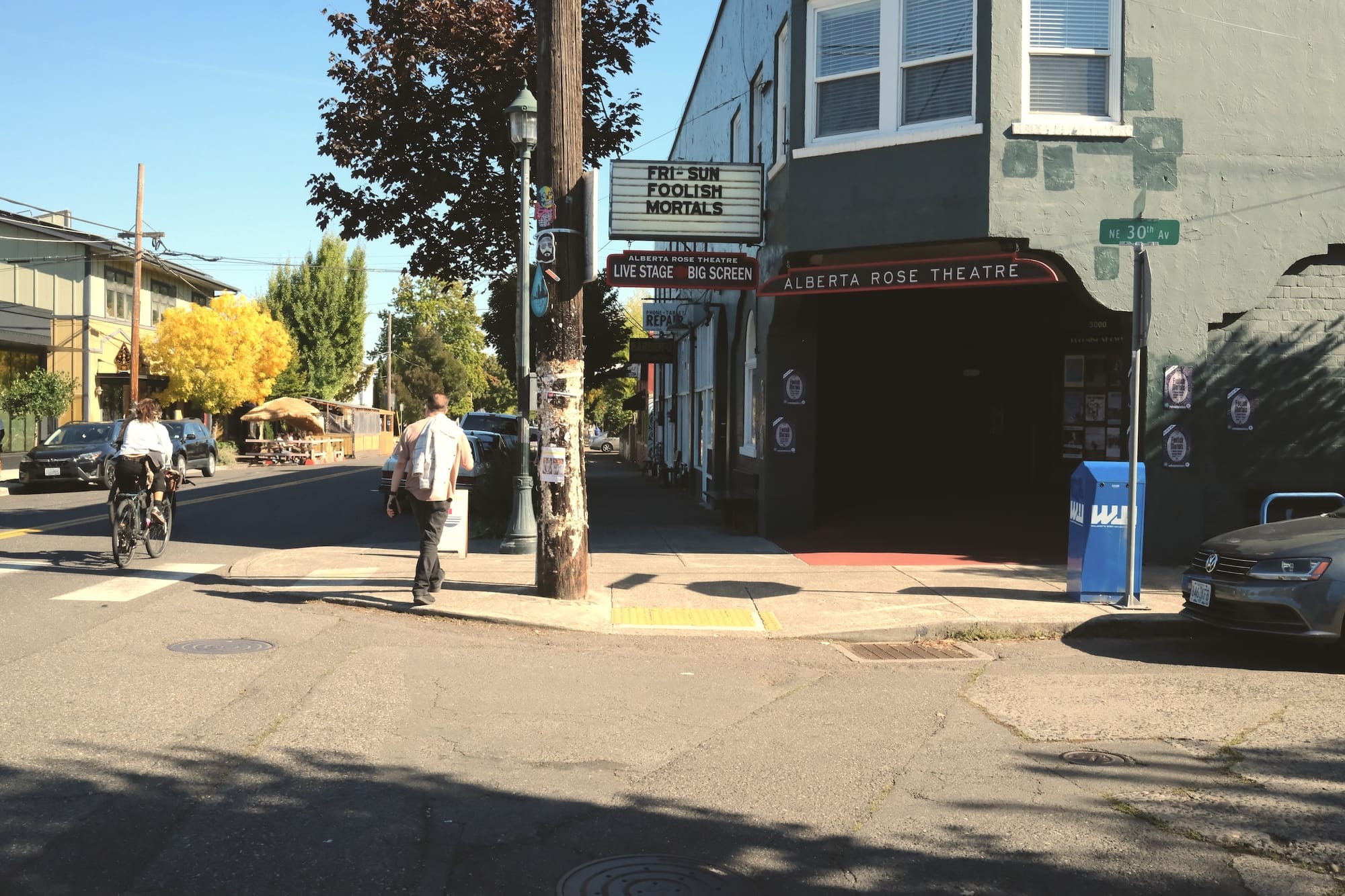 A cyclist zooms past the Alberta Rose Theatre, where the marquee sign reads "FRI-SUN FOOLISH MORTALS."