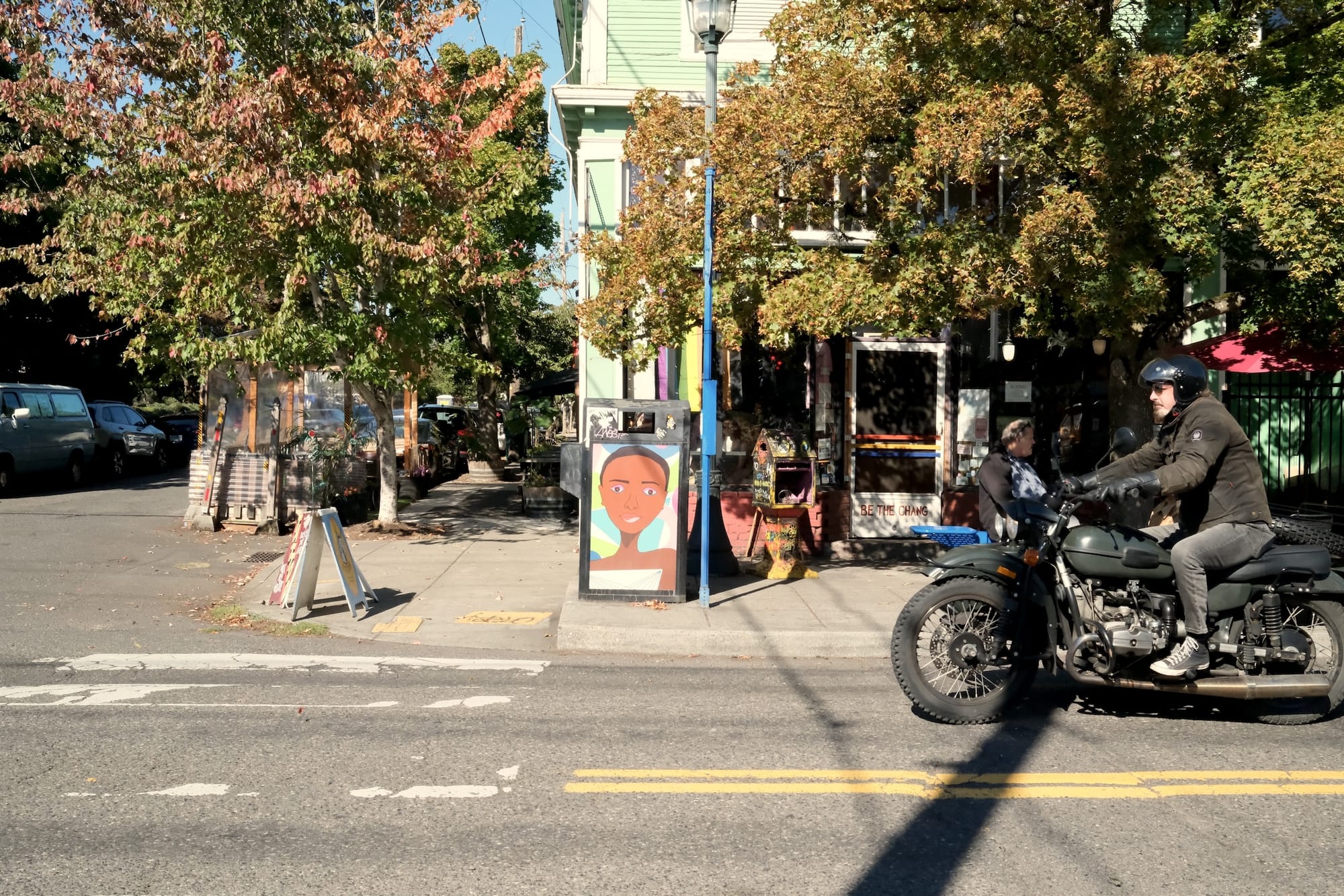 Faces adorn the trash cans in the Alberta Arts District. A motorcycle zooms by a coffee shop with a rainbow flag in the window. Leafy trees shade the streets.