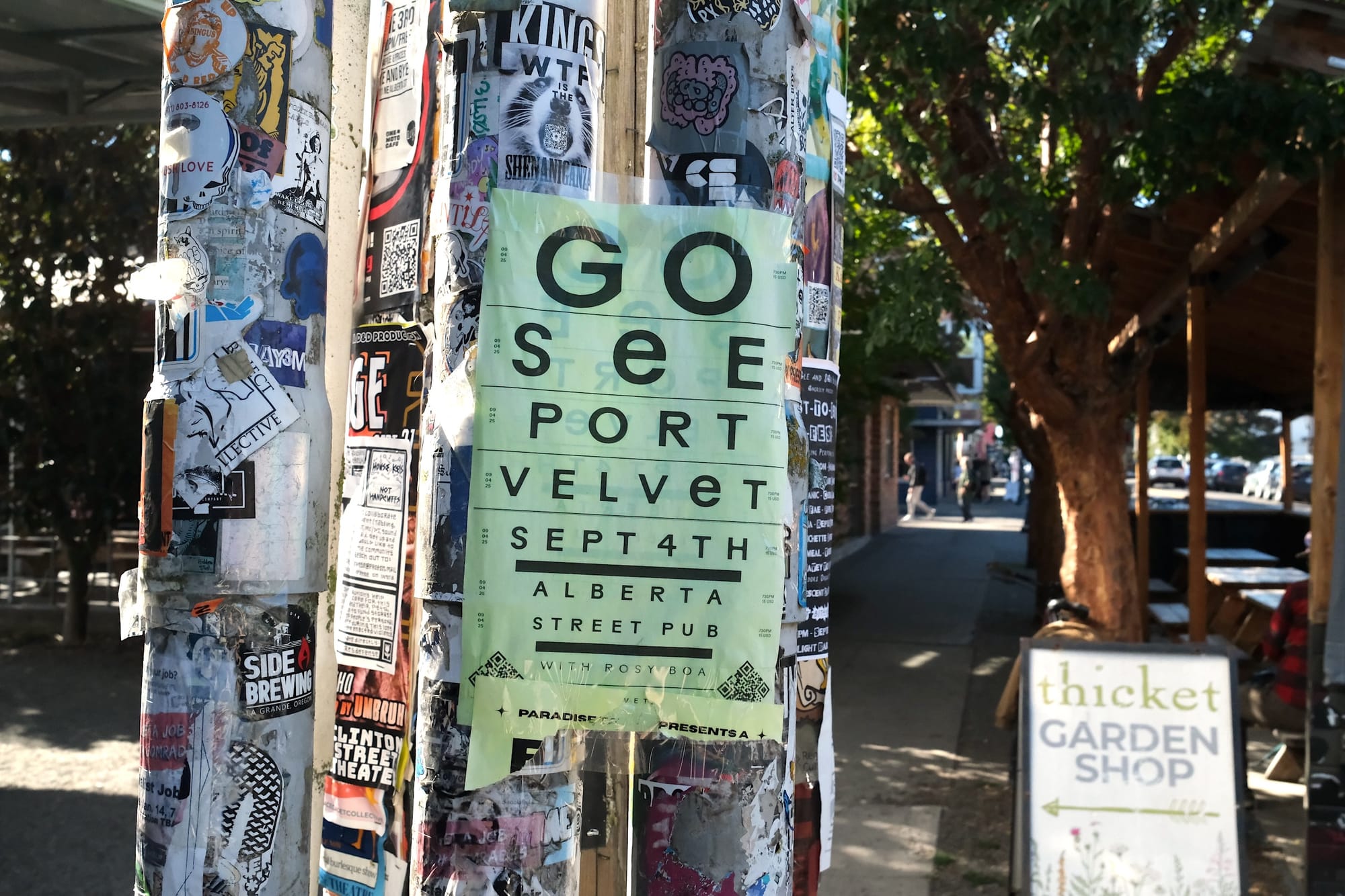 A poster resembling vision tests reads "Go See Port Velvet Sept 4th," while a sign board for a garden shop stands in the background.
