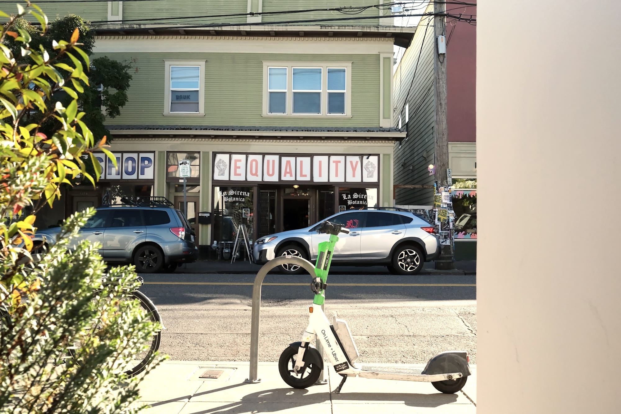 An e-scooter parks in front of a bike rack. Across the street, the storefront reads "EQUALITY" in capital letters and fist icons on both sides.