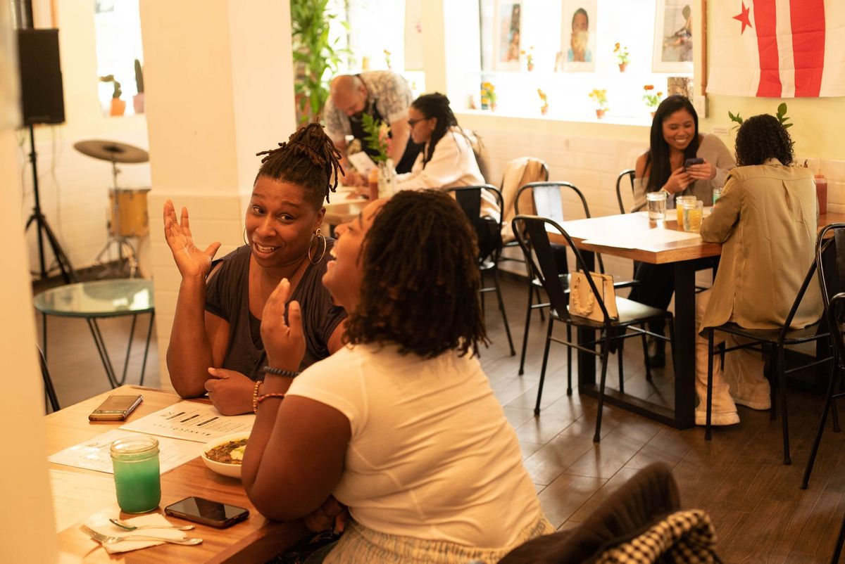 Tiara Darnell sits at a table with another diner, who laughs. Smiling customers fill tables in the background, along with a drum kit.