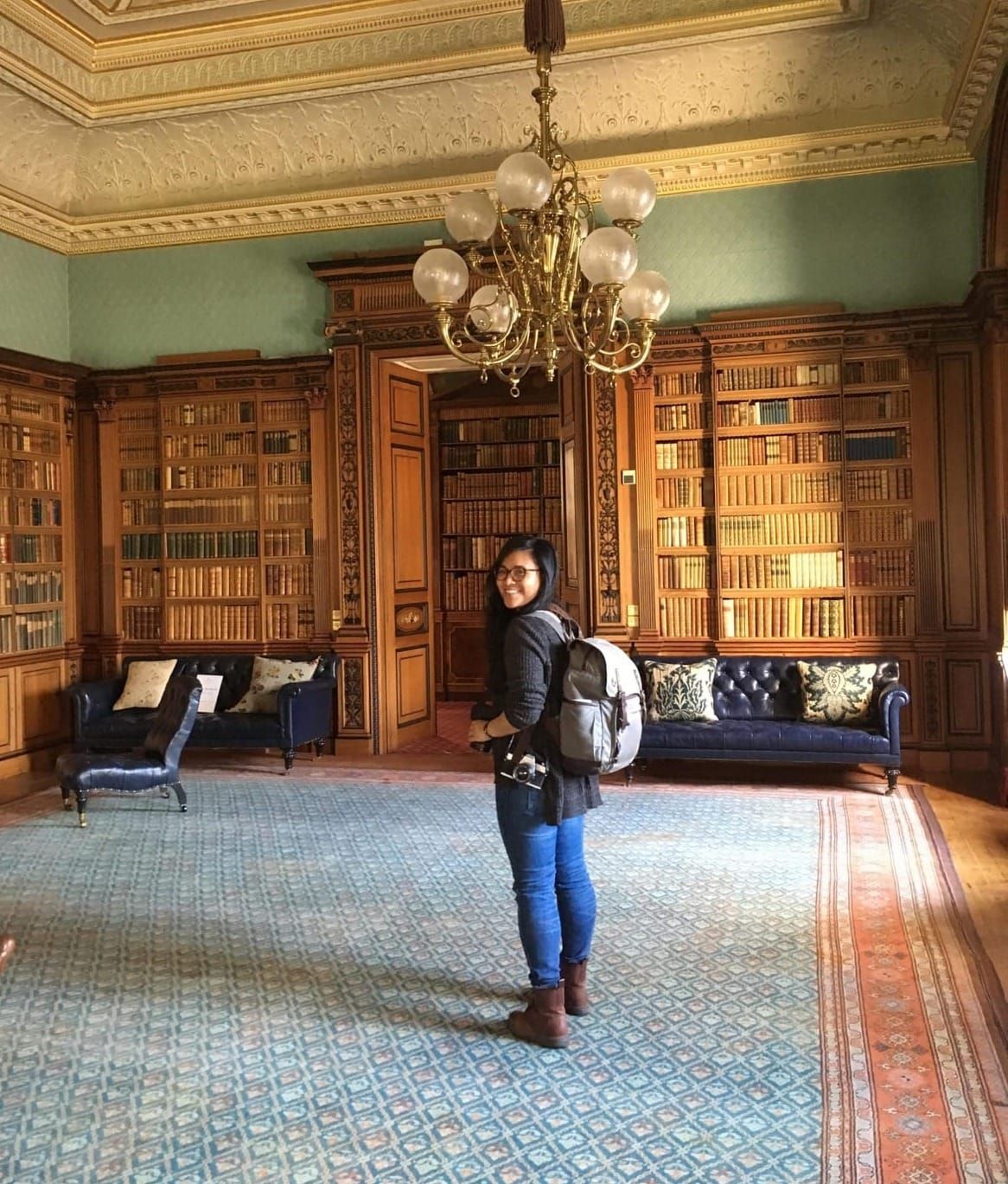 Photographer Celeste Noche stands in a grand room beneath a chandelier. She has a backpack on and a couple cameras around her neck. She's turned back, looking at the camera.