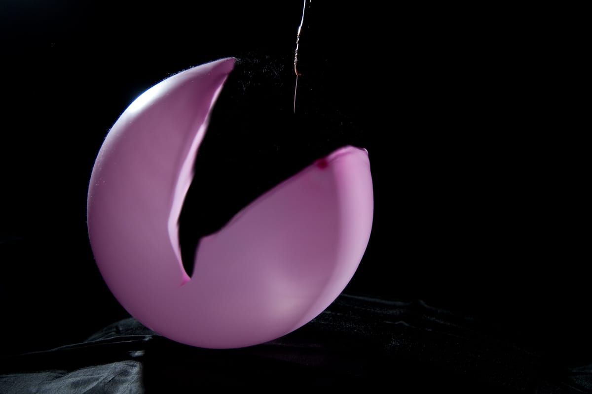 A high-speed photograph of a pink balloon at the moment of bursting, with its stretched membrane beginning to tear against a black background.