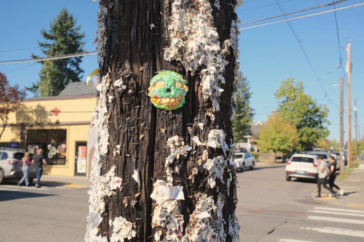 A little monster face screwed to a utility pole in Portland, Oregon, with a sunny street scene in the background and evergreen trees and blue skies in the distance.