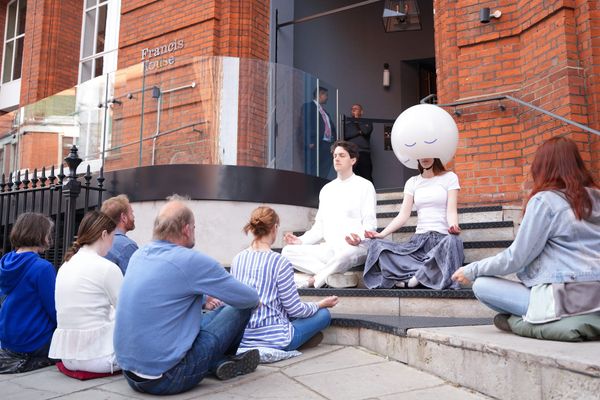 A group of people sit in meditation poses on the concrete steps outside a red brick building. One person wears a large white spherical helmet with a peaceful expression drawn on it.