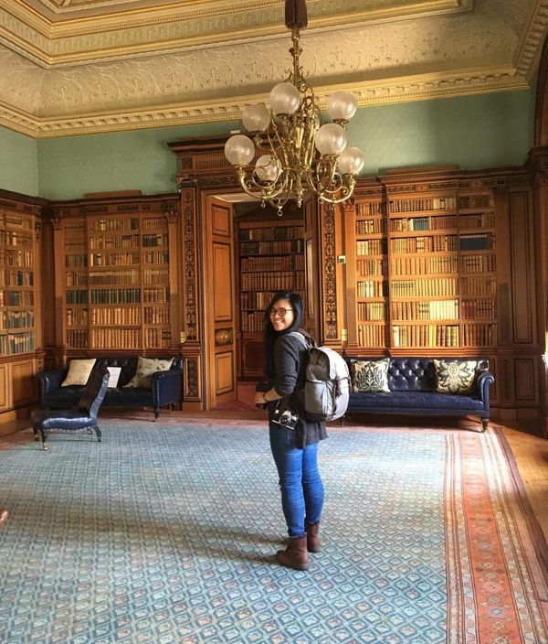 Photographer Celeste Noche stands in a grand room beneath a chandelier. She has a backpack on and a couple cameras around her neck. She's turned back, looking at the camera.