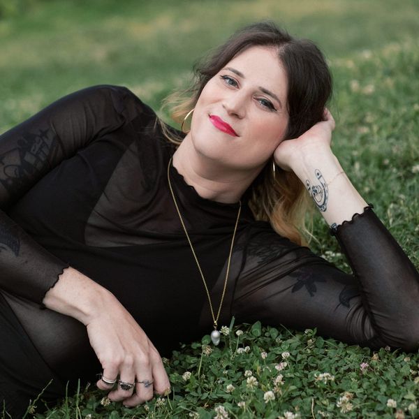 The artist lies in the grass, her head propped on one hand, the other hand rests in the cover, showing her three stylish rings.