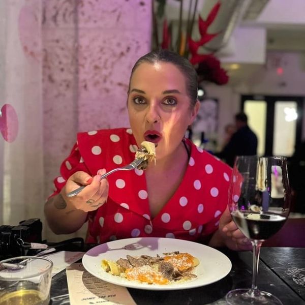 Alica Kennedy sits in a pink-dappled restaurant, wearing a polka-dot red dress and red lipstick, she opens her mouth and lifts a fork — a glass of red wine before her.