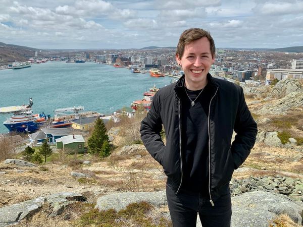 Paris Marx, dressed in all black and wearing a zip-up jacket, stands overlooking a bay behind him.