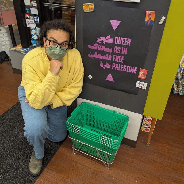 Alex Hanna, wearing a yellow sweater and blue jeans, kneels by a sign in a bookstore reading "queer as in free Palestine."