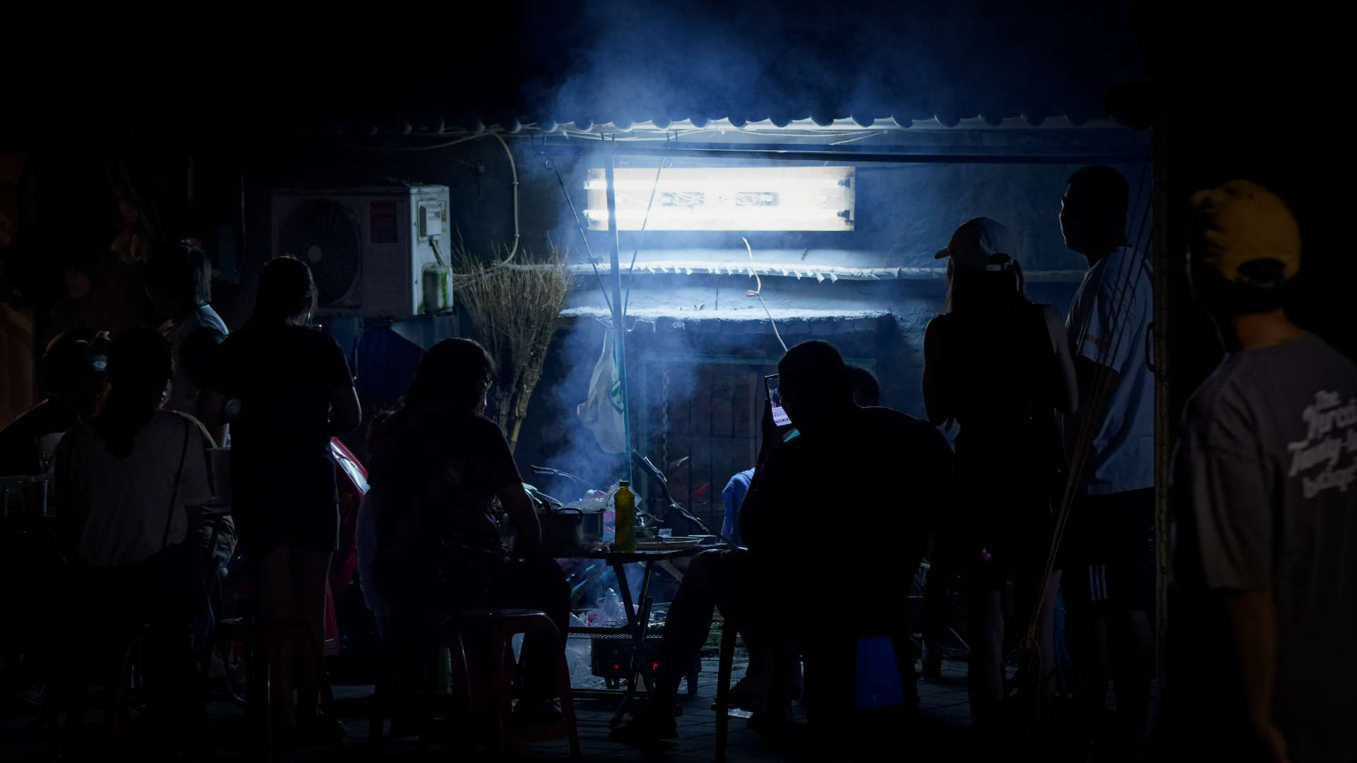 Silhouette of a group of people sitting around an outdoor barbeque at night, with smoke rising from the fire beneath the hot plate.