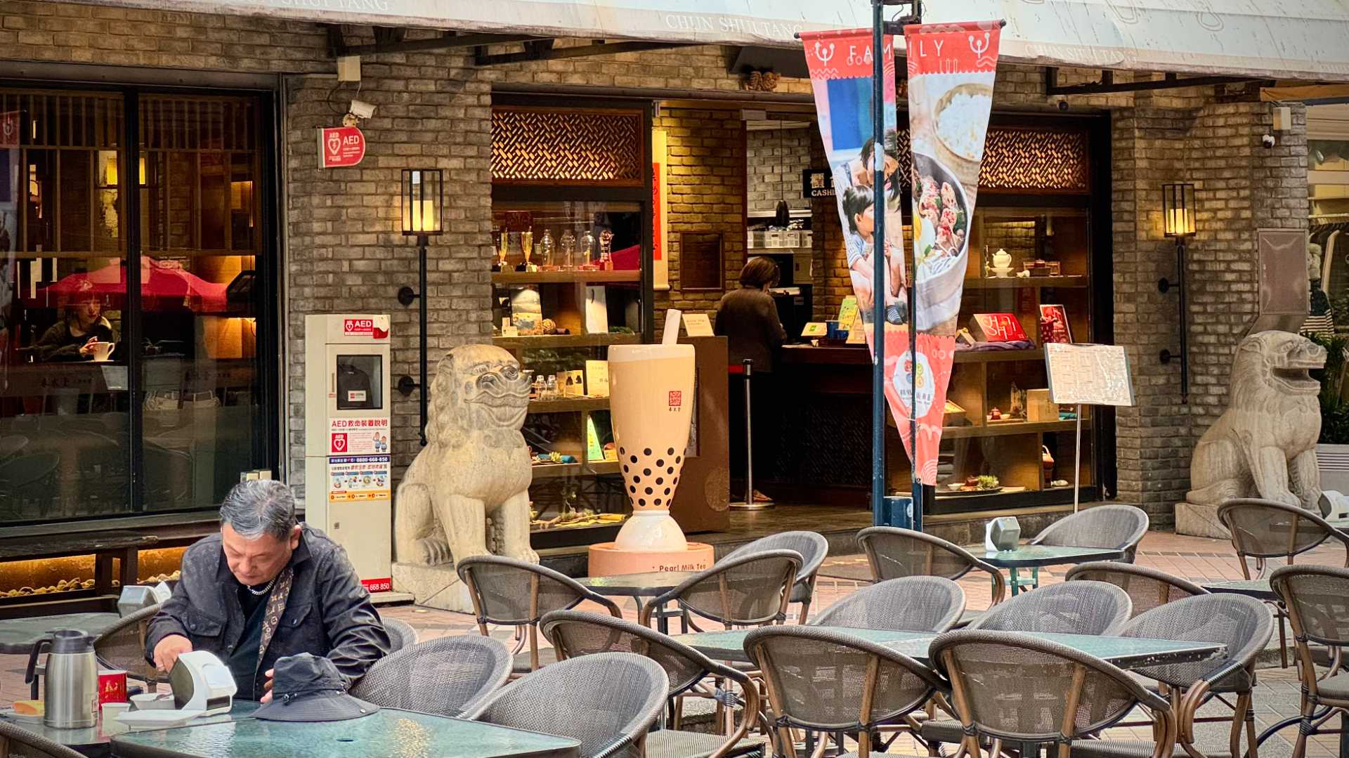 An elderly man sits alone drinking tea at one of many outdoor tables. Behind him is the entrance to a Chun Shui Tang teahouse, with stone lions either side of the door, plus a 1.5 meter-tall sculpture of a glass of bubble tea next to one of the lions.