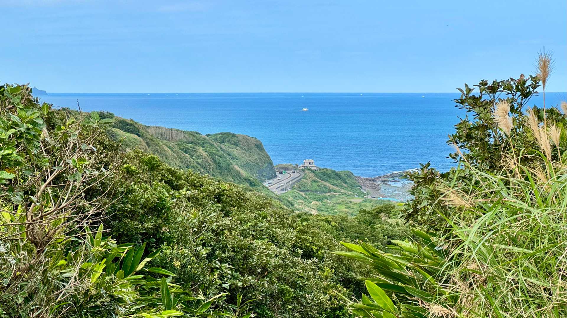 A view along a green bush-covered coastline. A wide road with car and truck traffic sweeps around a bend in the distance.