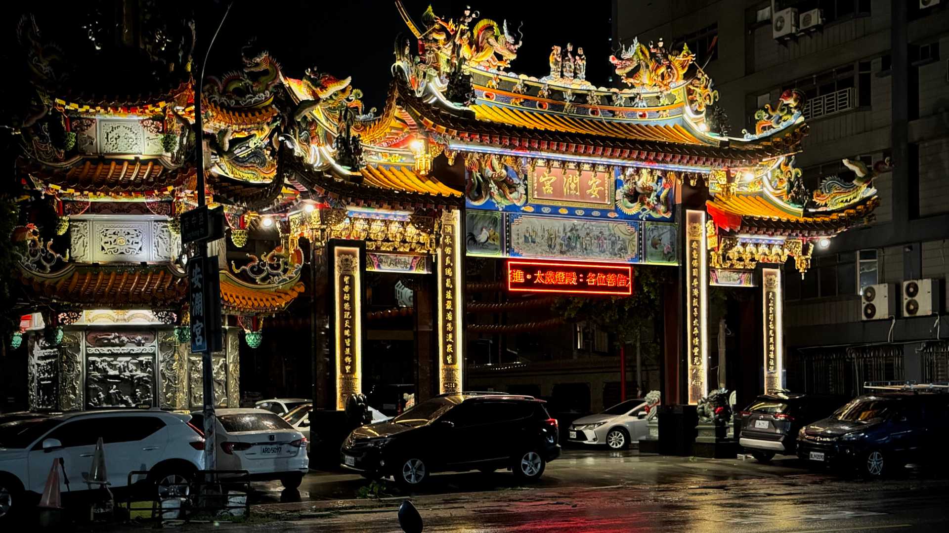An illuminated temple gate with a dozen or more cars parked on the forecourt. The colors are reflected on the wet tarmac.