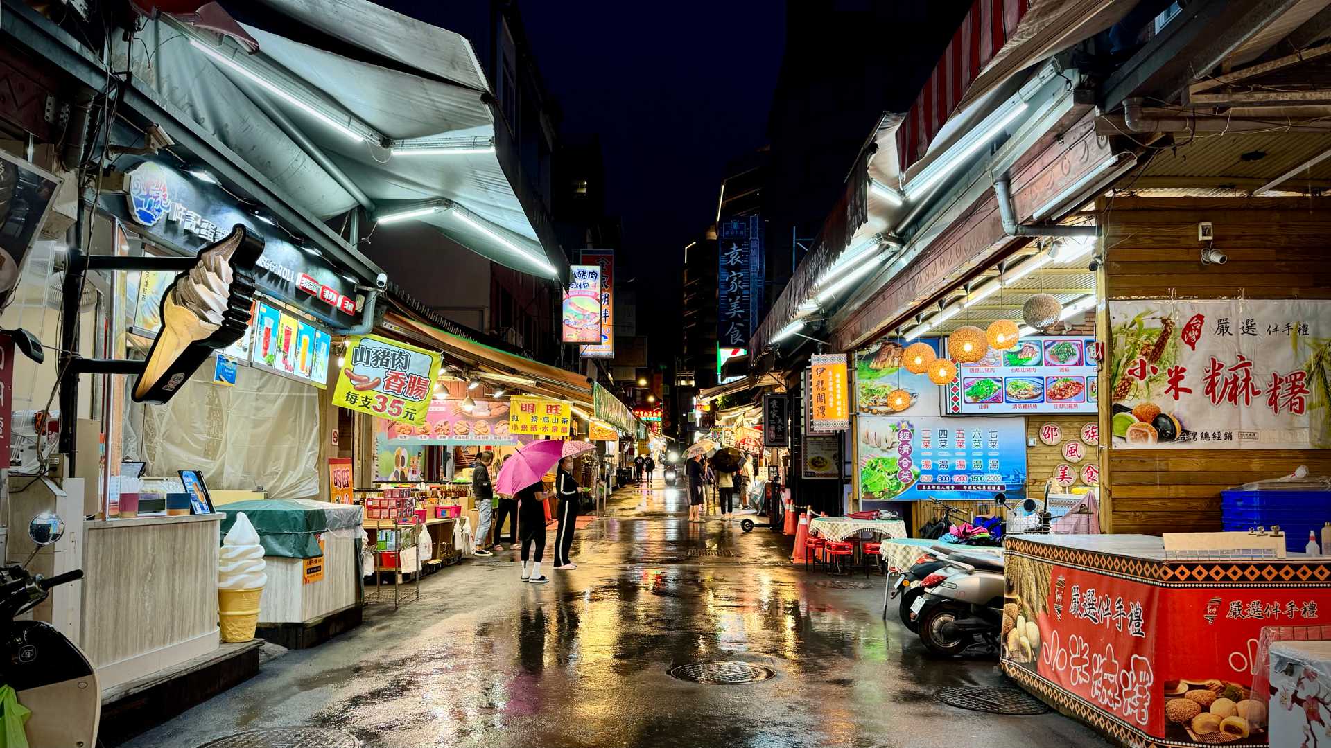 Wide-angle view of Ita Thao Night Market. There are only a few people walking about with umbrellas; the ground is wet and reflective because of the rain.