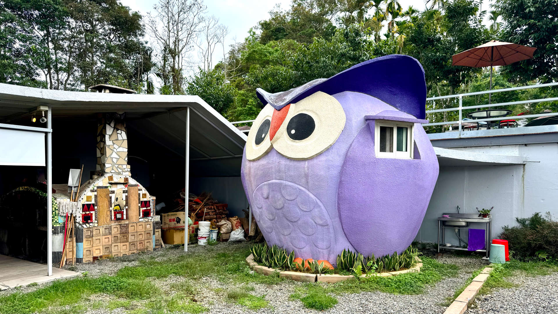 An outhouse building, approximately 2m in diameter, in the shape of a large cartoonish owl. An outdoor washbasin is visible behind it.