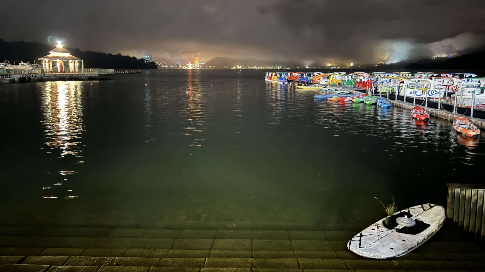 A photo of Sun Moon Lake take at night. There is a stand-up paddleboard resting against the shore in the foreground, with dozens of canoes and paddle-boats moored at a jetty in the background. Distant lights can be seen on the far side of the lake, perhaps 1km or 2km away.