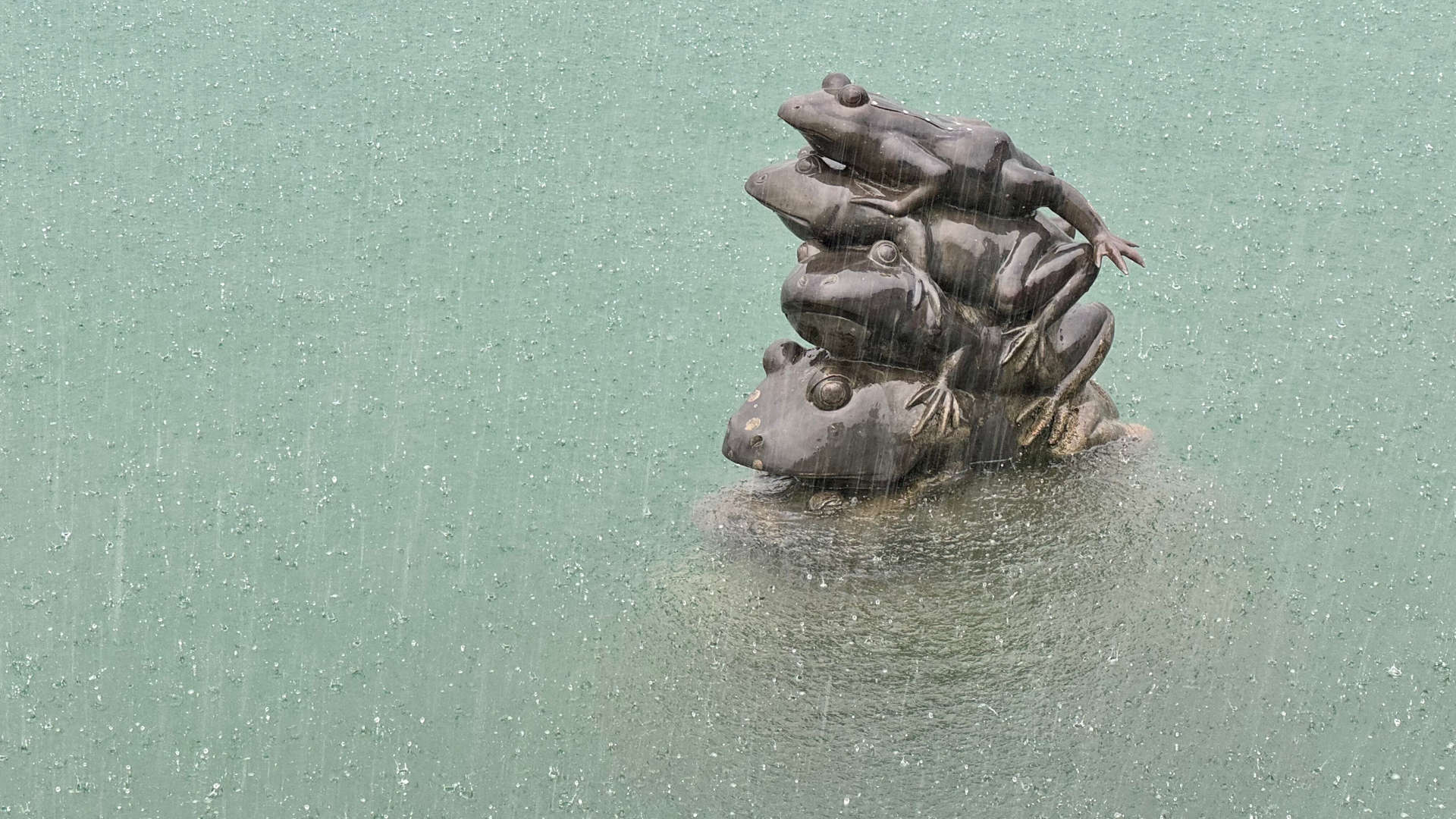 Heavy rain falls on a statue of stacked frogs rising out of the water. Four frogs are visible, stacked on top of one another.