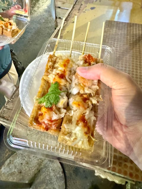 Point-of-view shot of a hand holding a plastic take-out container, which has two rectangular blocks of fried tofu inside. The fried tofu is topped with onions, cilantro, and sauces, with skewers protruding from one end.