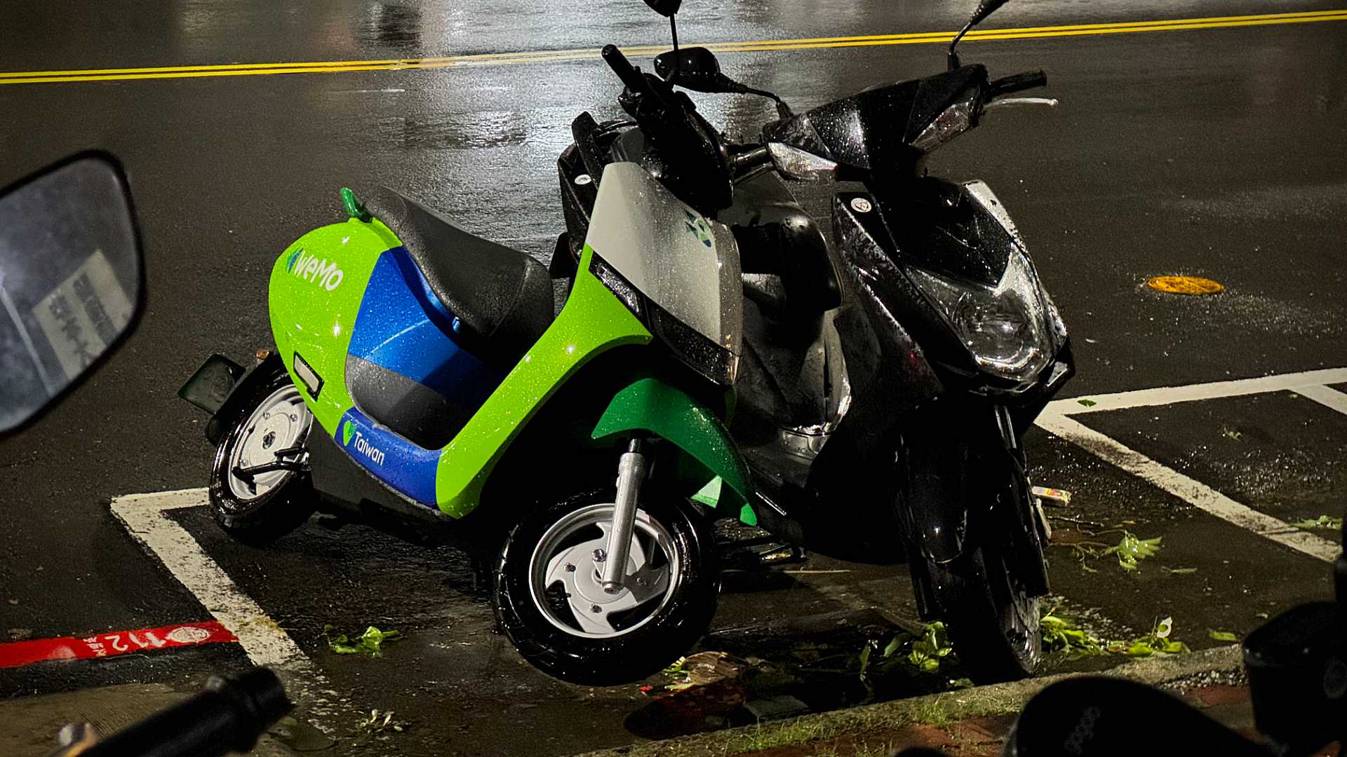A WeMo electric scooter on a lean, against a conventional petrol-powered motorcycle, after Typhoon Gaemi.