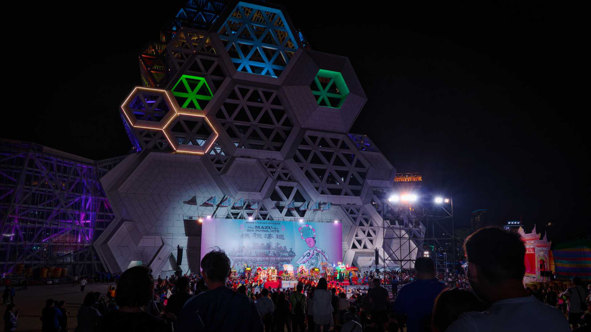 A crowd of thousands of people silhouetted in front of the Kaohsiung Music Center at night. In the distance, a group of perhaps two-dozen schoolchildren are performing in front a statues of various gods.