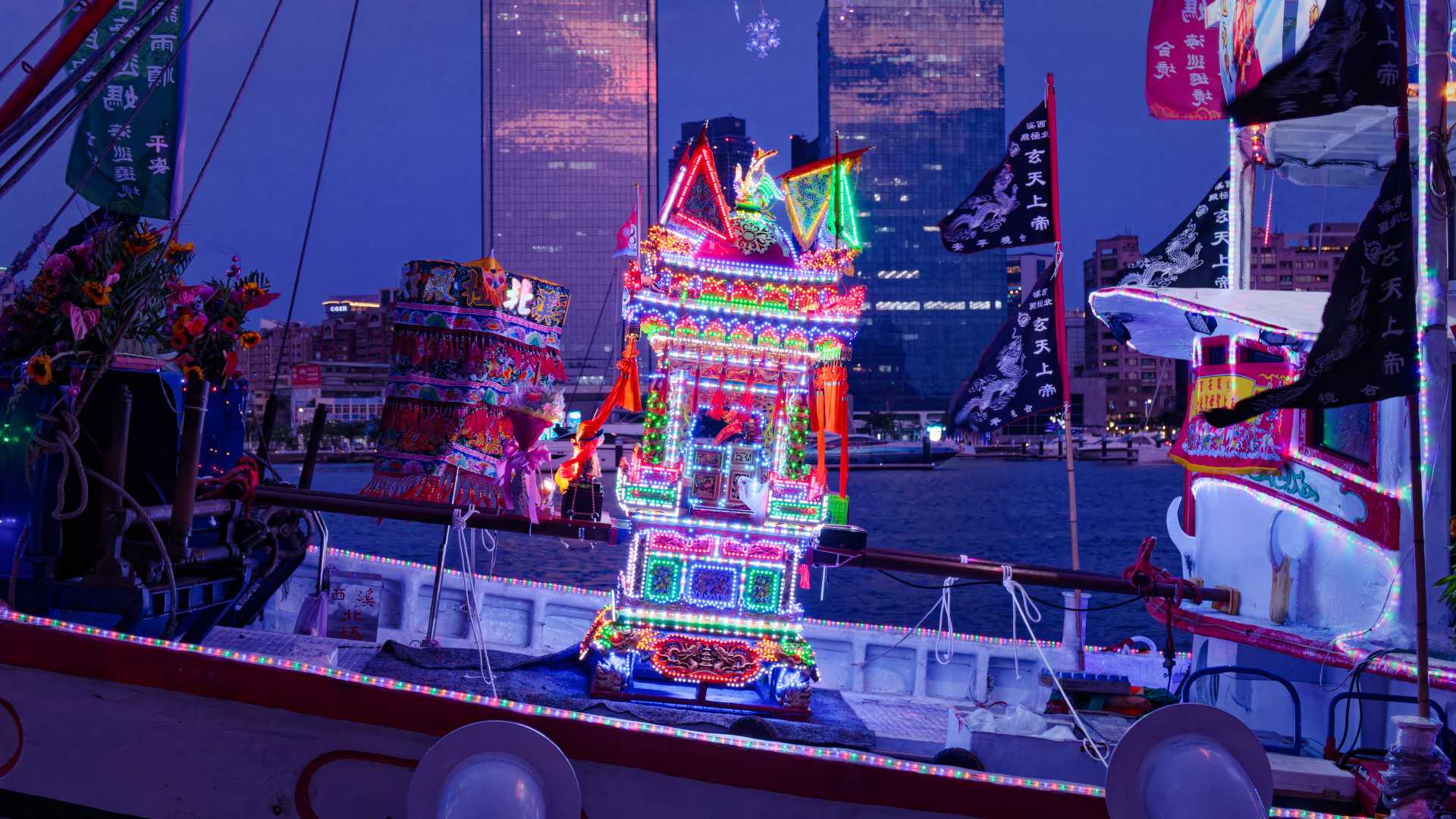 A brightly-lit and decorated shrine box on the forward deck of a Taiwanese fishing boat. It is just on dusk. Skyscrapers are visible it the distance across the Kaohsiung Harbor.