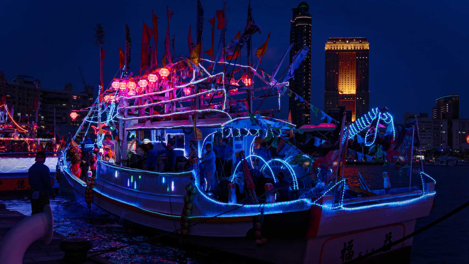 LED rope lights wrapped around equipment on the rear deck of a Taiwanese fishing boat. It's night, and skyscrapers can be seen in the distance across the harbor of Kaohsiung.
