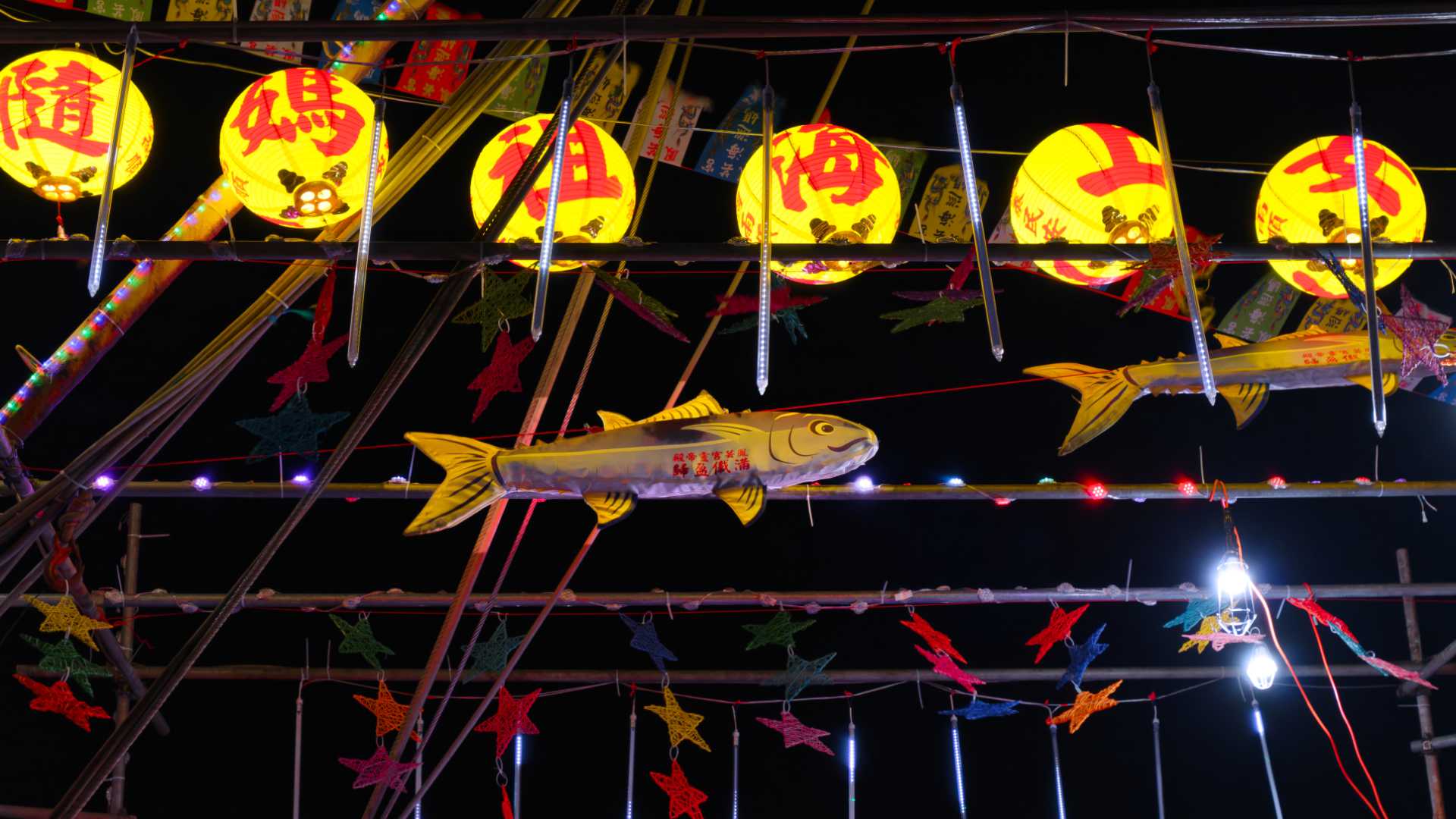 Inflatable fish and colorful woven star shapes hanging from the rigging of a Taiwanese fishing boat at night.