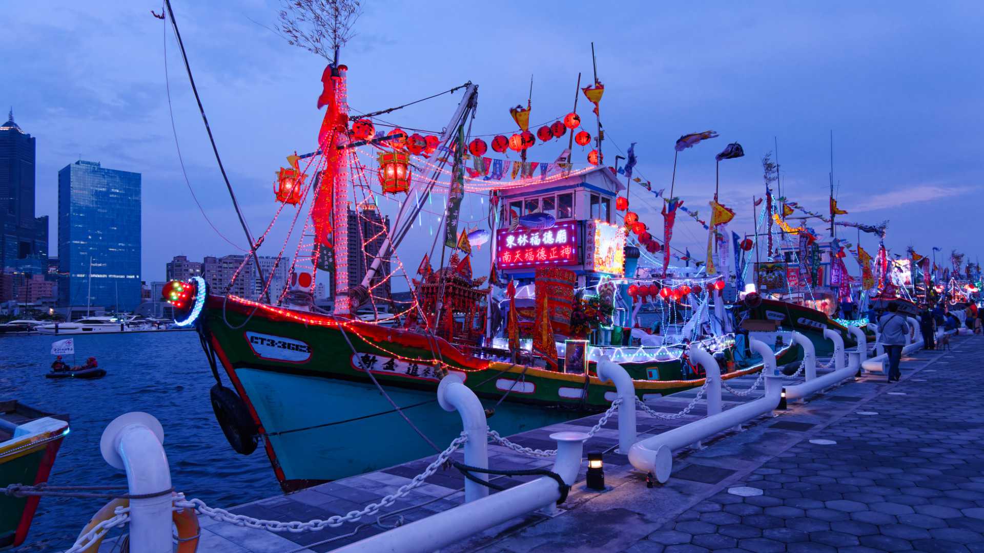A line-up of colorfully-decorated and illuminated fishing boats. The front one is decorated with dozens of hanging red lanterns.
