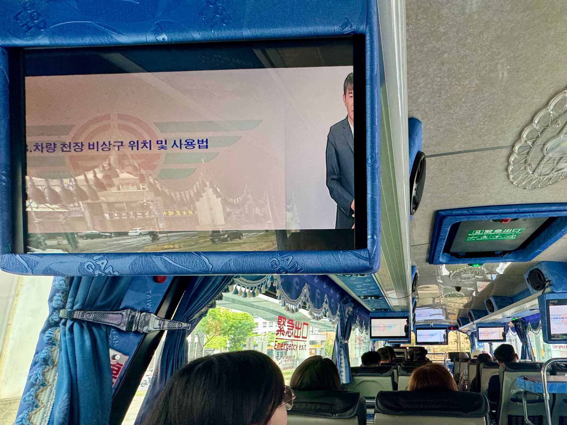 A ceiling-mounted TV screen inside a Taiwanese tour bus. The screen is showing a safety video with Korean characters on the screen, and a sign language interpreter half-cropped out of the extreme right edge.