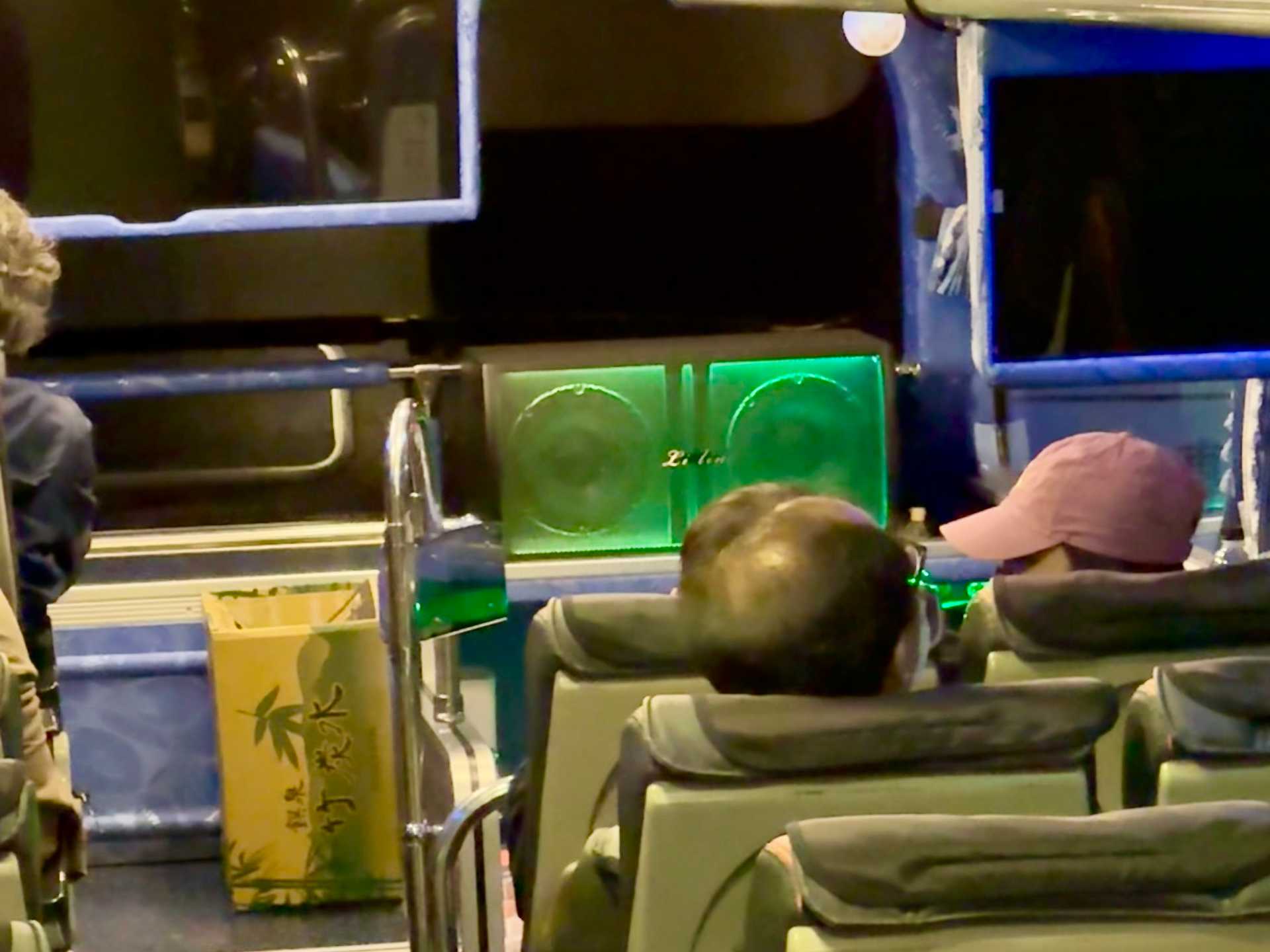 Zoomed photo of a large boombox-like speaker mounted next to a big-screen TV in front of the windshield at the front of the bus. The speaker is lit up in greenish-blue colors.