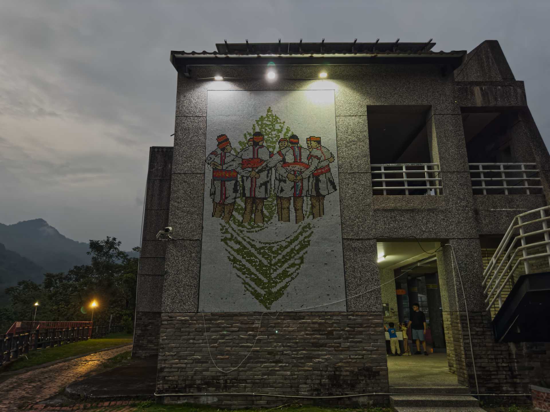 A two-story mosaic on the wall of a school building. The mosaic depicts a group approximately eight people in indigenous clothes standing arm-in-arm in a circle, with the shape of a cresent moon silhouetted against a leaf in the background.