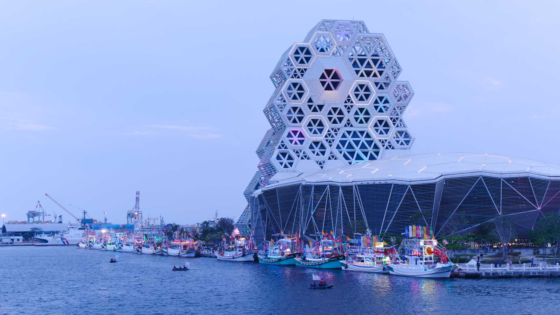 Around a dozen brightly-decorated fishing boats docked alongside Kaohsiung Music Center, just before dusk. It is a still, cloudy evening.