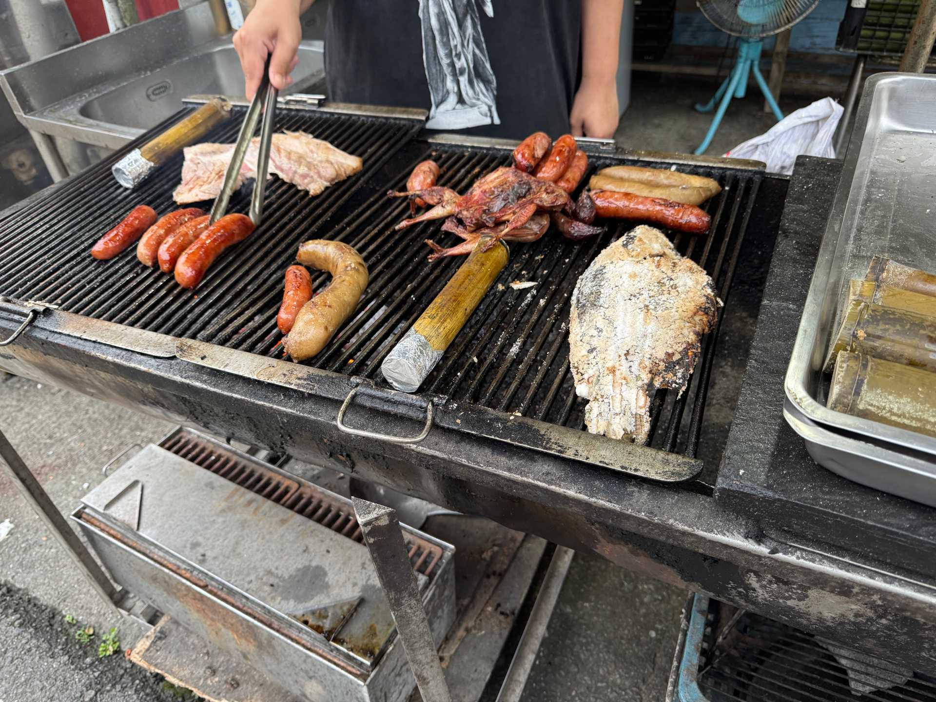 Close-up of various meats on a charcoal barbecue. Two sealed pieces of bamboo are also being roasted, in between a whole fish and a pigeon.