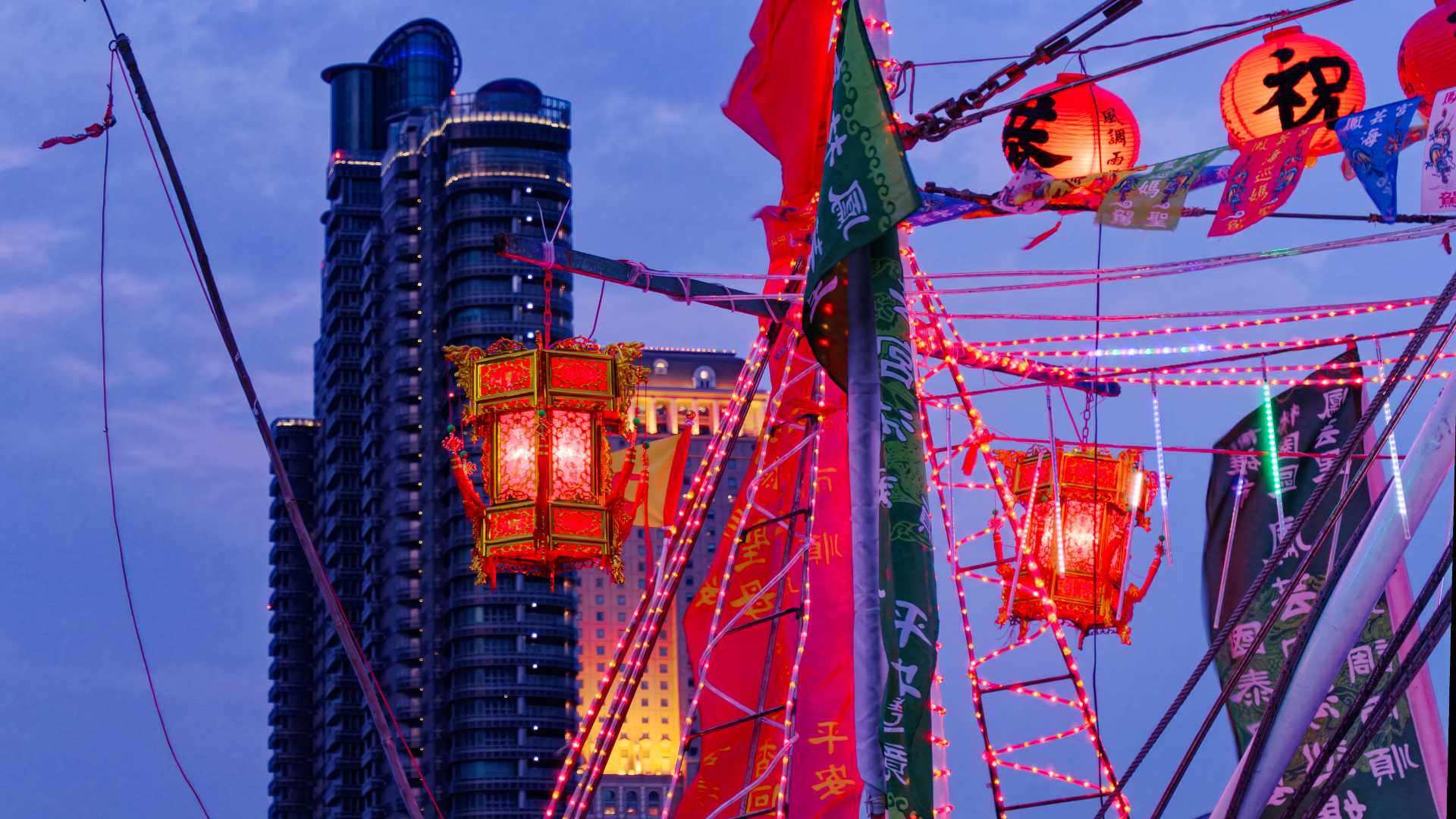 Close-up of two especially intricate red lanterns hanging from a Taiwan fishing boat. A skyscraper apartment complex is visible in the distance behind it.