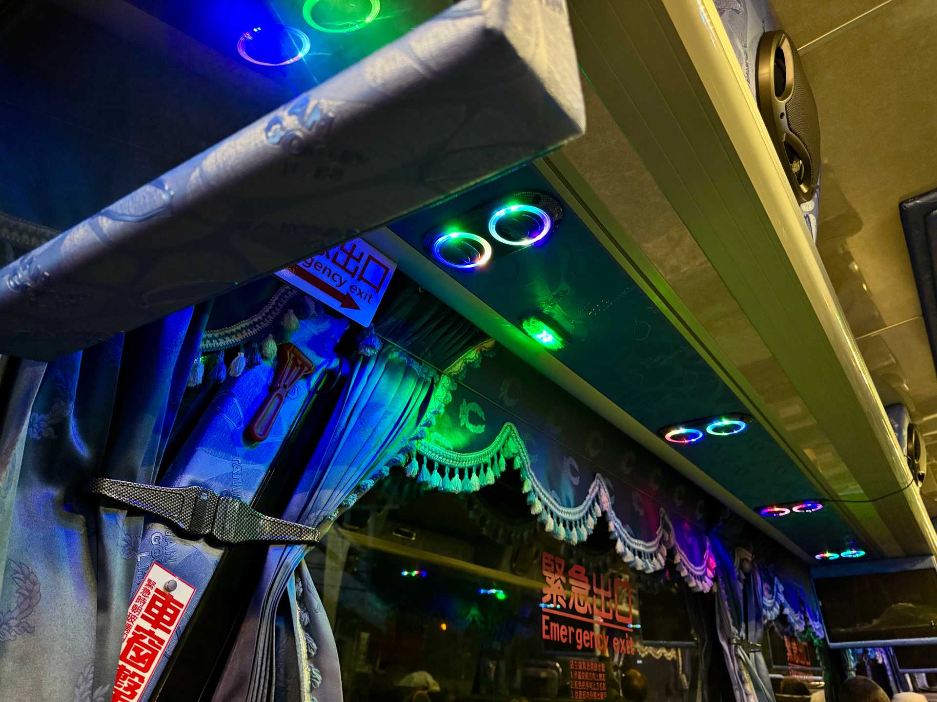 Interior of a Taiwan tour bus at night time. The air vents are ringed by colorful LED lights which also reflect off the frilly curtains and tassled valances above the windows.