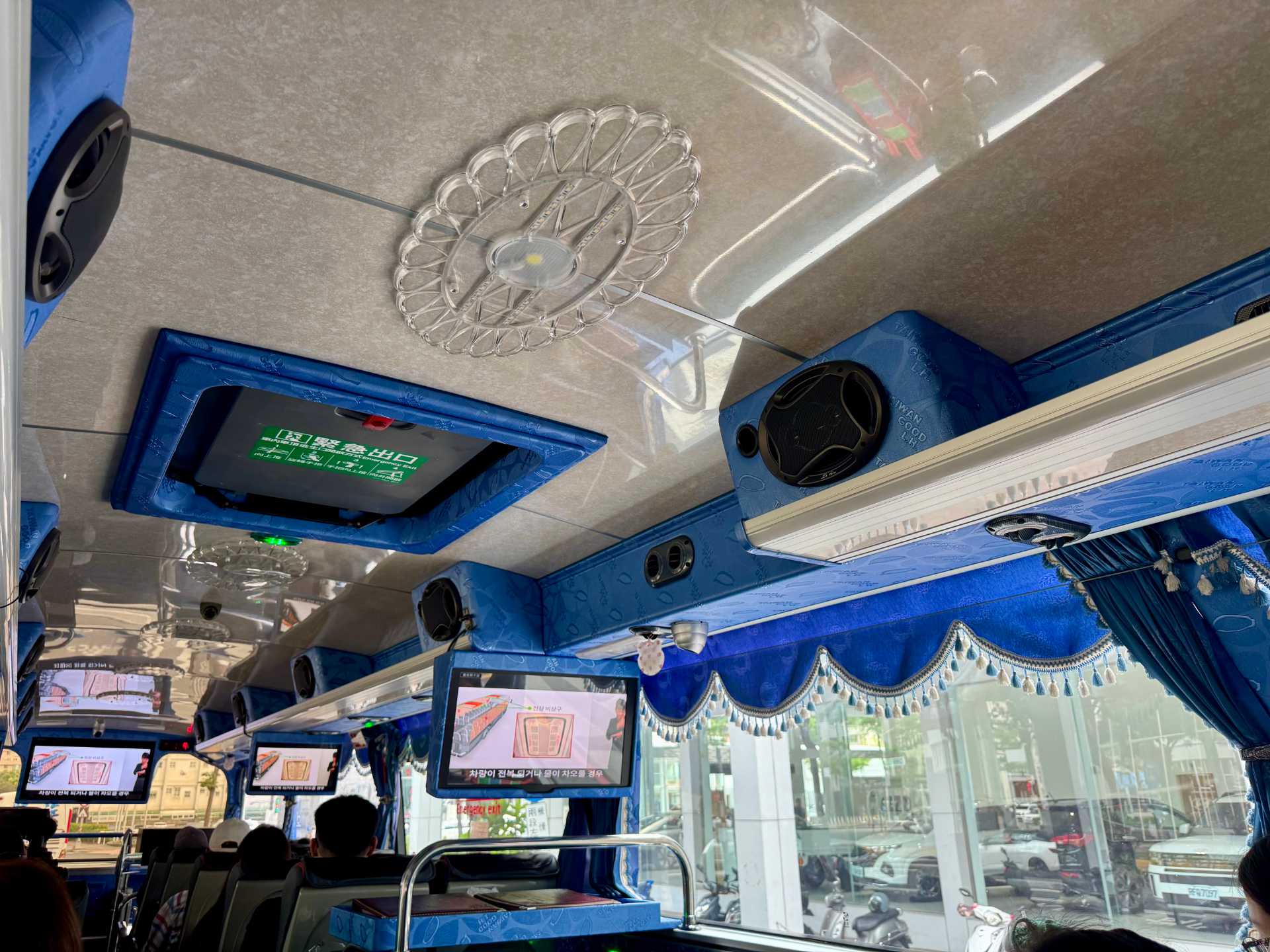 Daytime interior photo of a Taiwanese tour bus. The bus is decorated with rich blue curtains and valances, and dainty transparent plastic light fittings. The overall aesthetic is like something from the 1950s or 1960s.