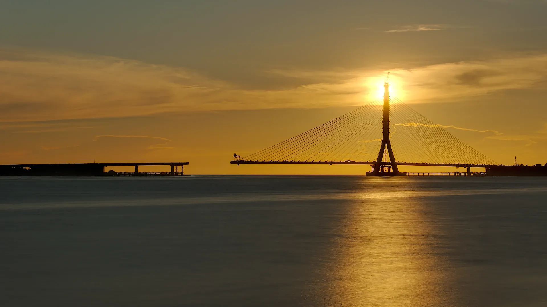 Danjiang Bridge, still under construction, photographed at sunset. The bridge is silhouetted against the sun, which is aligned with the bridge’s pylon.