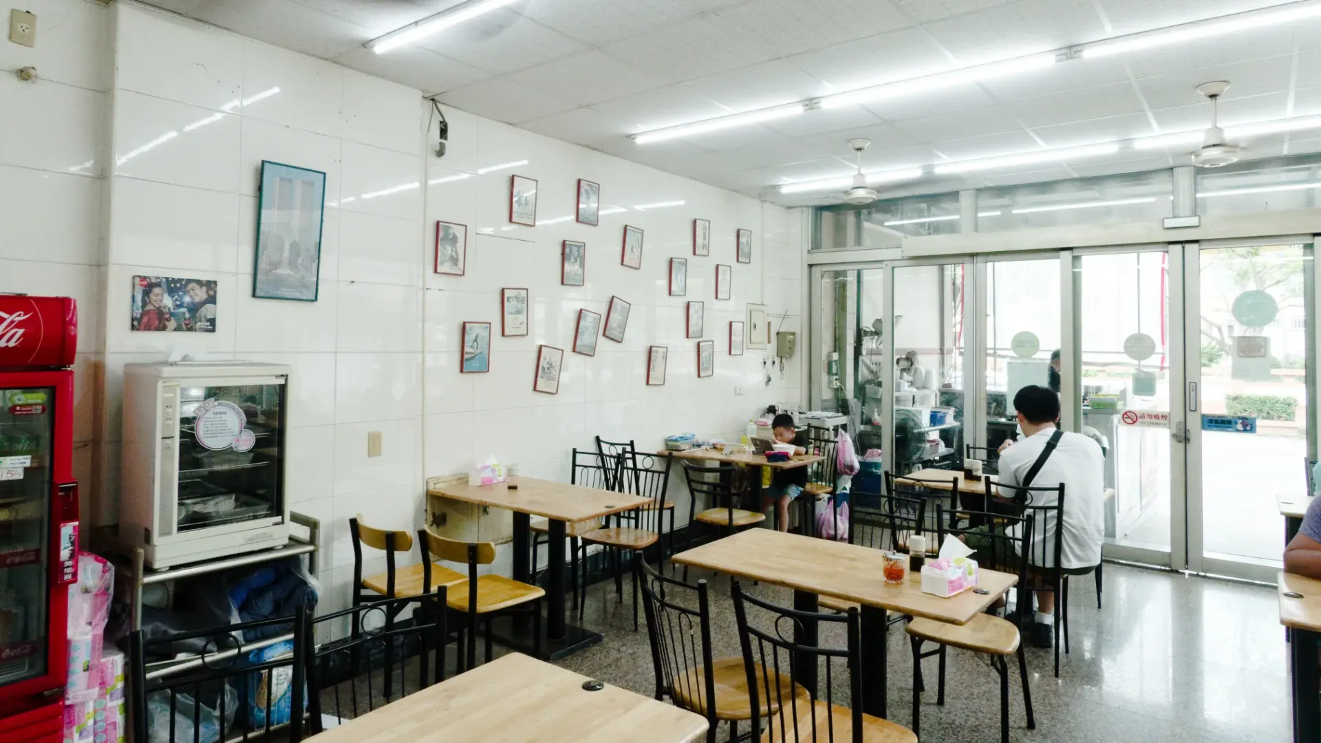 Interior of Jiaxiang Beef Noodle Restaurant. There are three customers at different tables. An array of framed posters, at haphazard angles, is visible on the far wall.
