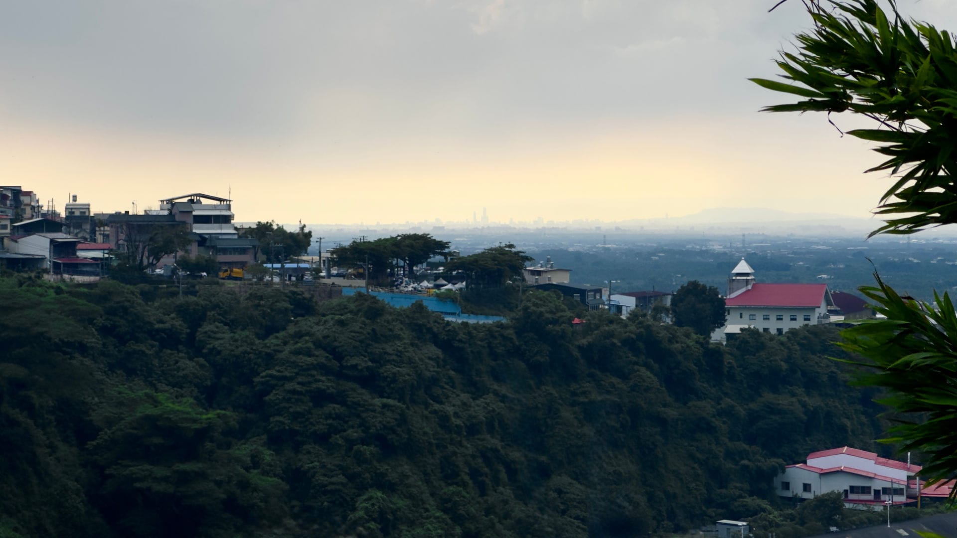 A zoomed-in view looking over a small village in the foreground, and the plains of Pingtung County, to the silhouettes of distance skyscrapers in Kaohsiung City, Taiwan.