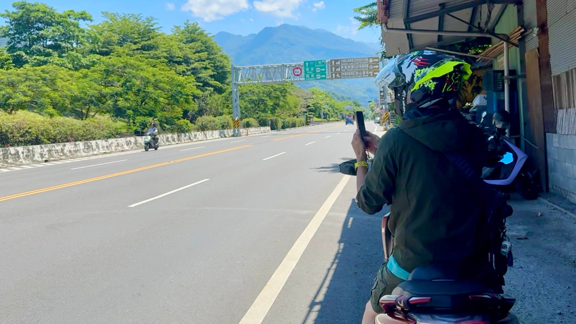 A four-lane rural road with mountains in the distance. In the foreground another scooter rider has stopped, and is using their phone to photograph an other scooter rider on the far side of the road.