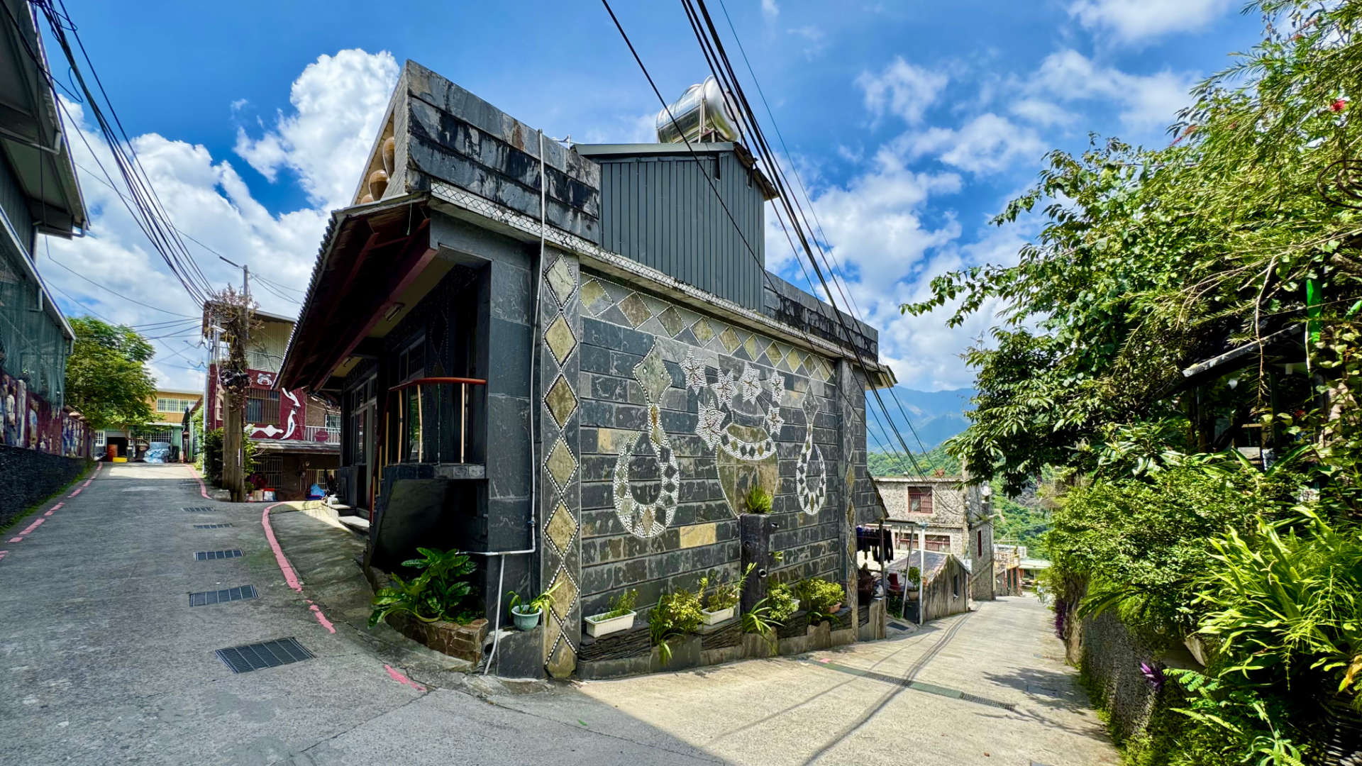 A concrete-and-stone house on the corner of two narrow streets in Wutai Township, Taiwan. One wall of the house is decorated with Rukai motifs depicting two snakes and a clay pot filled with flowers.