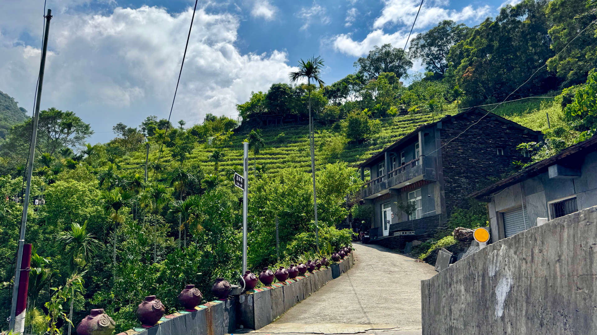 A narrow road leading up a steep hill in Wutai Township, Taiwan. The side of the road is lined with decorative clay pots. Further uphill, a two-story house is clad in slate. There are tiered agricultural terraces on the hillside beyond.